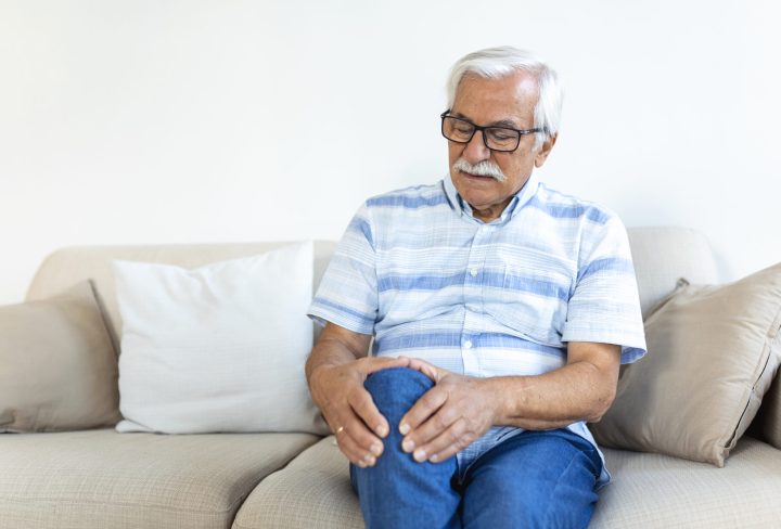 Elderly man sitting on a sofa at home and touching his painful knee. people, health care and problem concept - unhappy senior man suffering from knee ache at home